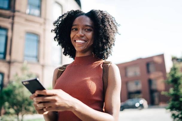 Smiling content African American curly haired woman in sleeveless casual shirt texting on mobile phone in street in summer and looking at camera - Foto, Imagen