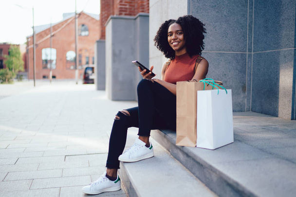Happy content white toothed African American curly haired woman sitting on stairs with paper bags in sleeveless casual shirt texting on mobile phone in street in summer - Foto, imagen