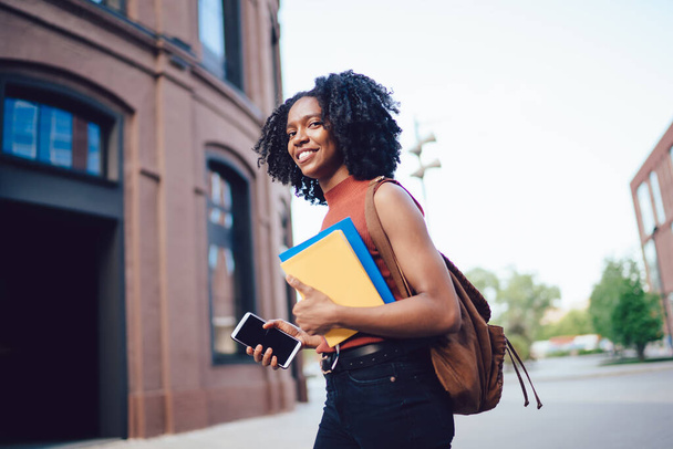 Happy content African American curly haired woman in sleeveless casual shirt  walking with smartphone and books and backpack in hand in street in summe - 写真・画像