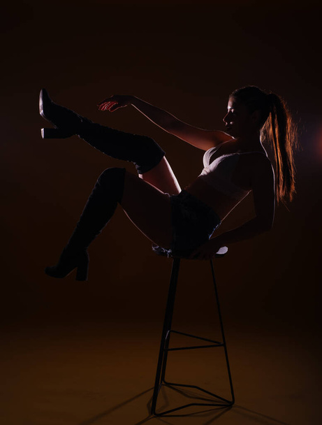 Высокий контрастный силуэт фотографии сексуальной и страстной молодой женщины, сидящей на стуле и позирующей с поднятыми ногами - Фото, изображение