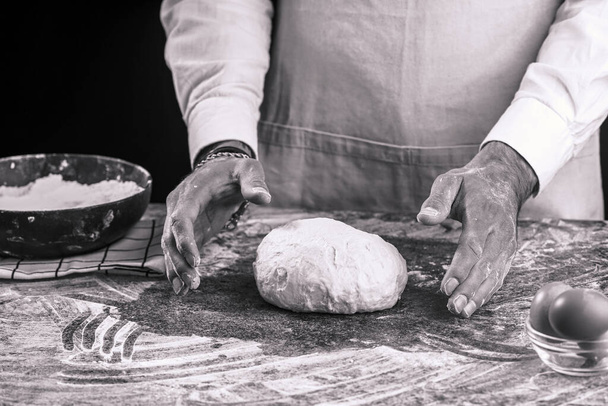 A male chef baker in a white shirt and grey apron kneading dough on a dark kitchen table, black and white - Foto, Imagem