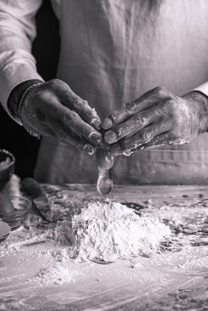 A male chef cook in a white shirt and grey apron breaking an egg into a heap of flour, black and white - Fotografie, Obrázek