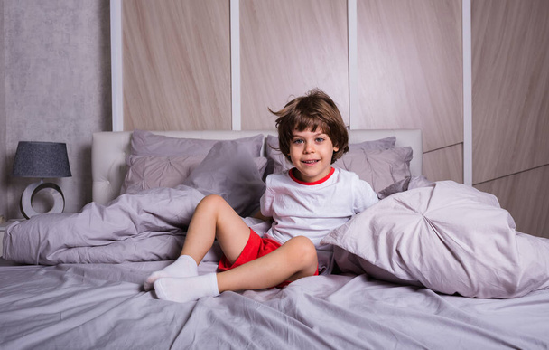 Pijamalı küçük mutlu çocuk yatak odasında yatakta oturuyor. - Fotoğraf, Görsel