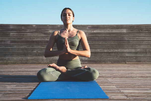 Slim peaceful ethnic female yogi sitting in lotus position with folded hands and closed eyes on mat on wooden sidewalk - Φωτογραφία, εικόνα