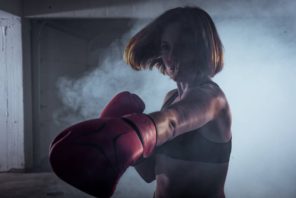 Sportowa dziewczyna z konkurencyjną postawą walki robi ćwiczenia bokserskie i robi bezpośrednie trafienie na zadymionym tle - Zdjęcie, obraz
