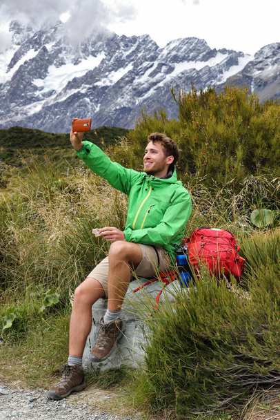New Zealand kiwi tramper taking selfie phone picture during hike on Hooker Valley track trail in Mt Cook. Summer hiker eating lunch break during hiking. Happy man taking photos - Foto, immagini