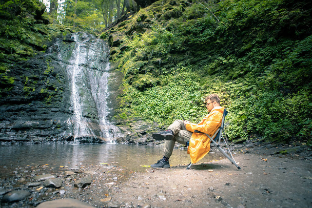 homem sentado em cadeira portátil na frente da cachoeira na floresta leitura livro cópia espaço - Foto, Imagem