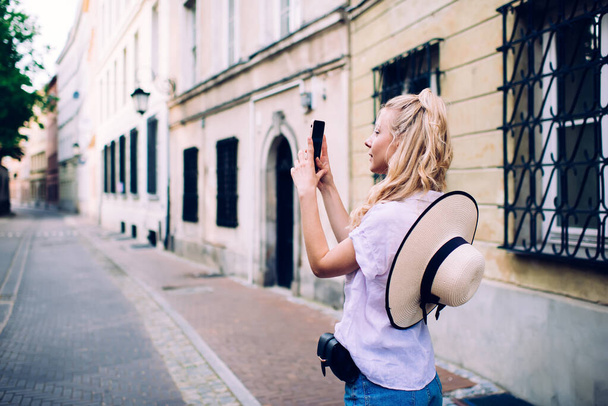 Side view of casual woman with blond hair in white blouse taking photo on building standing on old narrow empty street - Foto, immagini