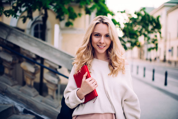 Confident romantic young blonde female graduate in casual wear holding red notebook while standing on street and looking at camera  - Фото, изображение