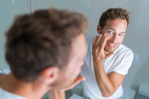 Man putting skincare facial treatment cream on face. Anti-aging skin care product. Male beauty morning routine at home lifestyle. Guy looking in bathroom mirror applying moisturizer under eyes - Fotó, kép