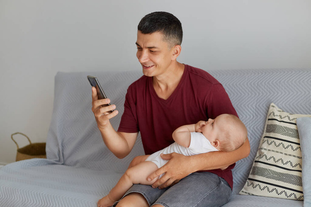 Portrait of young adult Caucasian man wearing maroon casual style t shirt with cute child in white attire, father holding his baby while using his phone while sitting on sofa in light room. - Fotó, kép