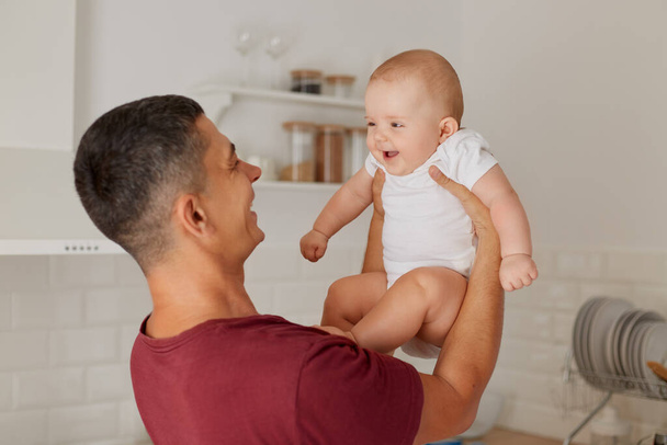 Portrait of happy positive father holding charming baby girl wearing white t shirt in hands, excited child likes spend time with daddy, family posing in light room. - Φωτογραφία, εικόνα