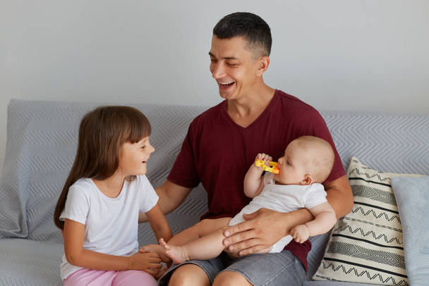 Indoor shot of happy family, smiling father playing with two daughters on sofa, cute baby biting toy, dad spending time with charming children at home. - 写真・画像