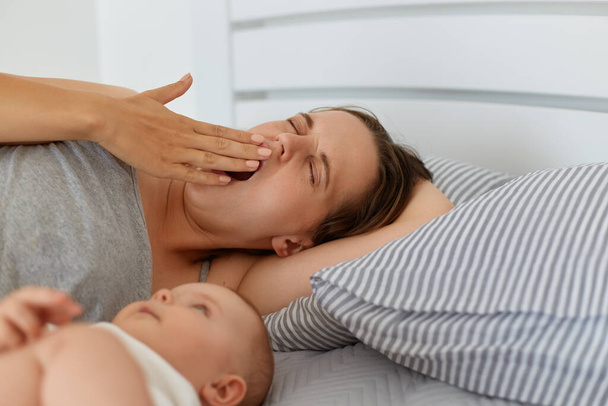 Indoor portrait of tired mother lying in bed with little baby girl or boy, yawning, covering mouth with palm, looks sleepy, having lack of energy and sleepless nights. - Foto, Bild