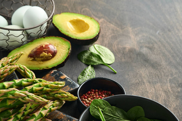 Bunch of fresh green organic asparagus spinach, avocado, chicken eggs and pepper seasoning on black old wooden background, top view. Food cooking background with copy space. Mock up. - 写真・画像