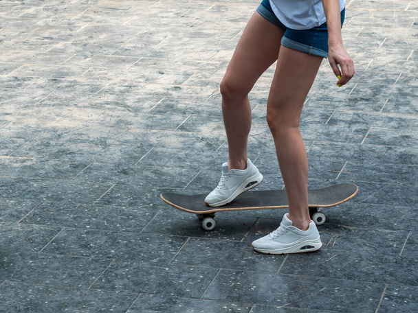 Close up photo of cool young girl with beautiful legs in white sneakers riding skateboard in shiny summer day . Outdoor activities. Street culture. - Fotó, kép