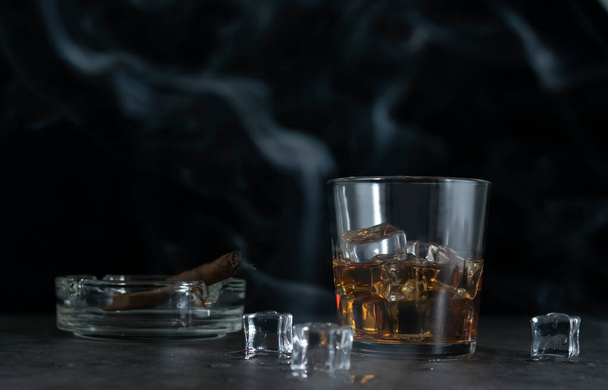 A glass of whiskey and ice cubes in clouds of blue smoke on a dark background. An ashtray with a cigarette in the background - Foto, afbeelding
