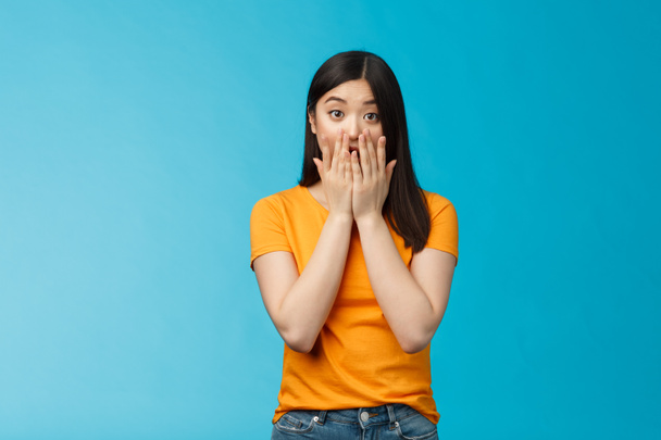 Shocked speechless stunned asian girl stare camera cover mouth palms, raise eyebrows gasping astonished, feel pitty sorry hearing bad news, stand surprised blue background wear yellow t-shirt - Fotografie, Obrázek