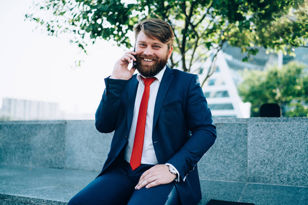 Joyful smiling bearded male in elegant suit chatting on mobile phone while sitting on tiled fence in park of modern city - Foto, Bild