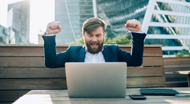 Happy bearded businessman with raised hands in formal wear looking at laptop celebrating successful startup sitting at table in park of modern city - Photo, Image
