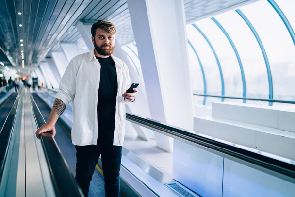 Half length portrait of confident male dressed in casual apparel looking at camera while waiting for email answer on mobile gadget, millennial Caucasian man with cellular phone standing at escalator - Фото, зображення