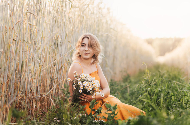 Mladá krásná dívka sedí na pšeničném poli a drží v rukou kytici sedmikrásek. - Fotografie, Obrázek