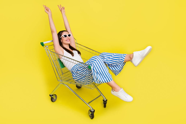 Portrait of attractive cheerful girl sitting in cart having fun showing v-sign isolated over bright yellow color background - Fotó, kép