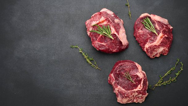 raw piece of beef ribeye with rosemary, thyme on a black table, top view, copy space - Foto, Bild