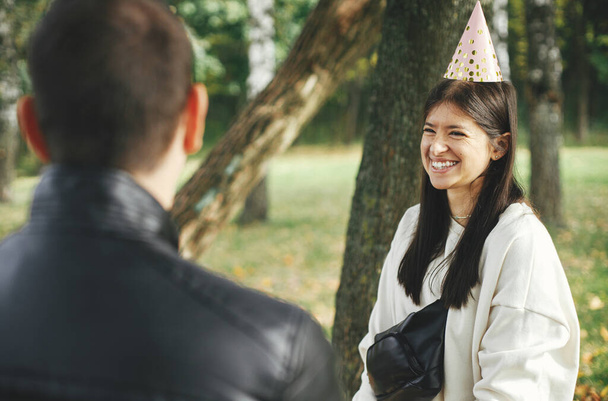 Стильная молодая счастливая женщина в шляпе, празднующая день рождения с семьей и друзьями на пикнике под открытым небом. Мужчина поздравляет подругу с днем рождения. Семейное собрание в парке - Фото, изображение