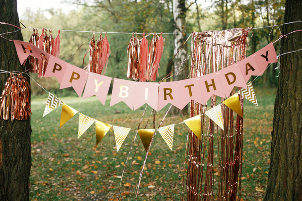 Stylish happy birthday garland hanging in park. Modern rose gold decor, tassel garland and happy birthday banner hanging on trees picnic party. Birthday celebration outdoor - Photo, Image