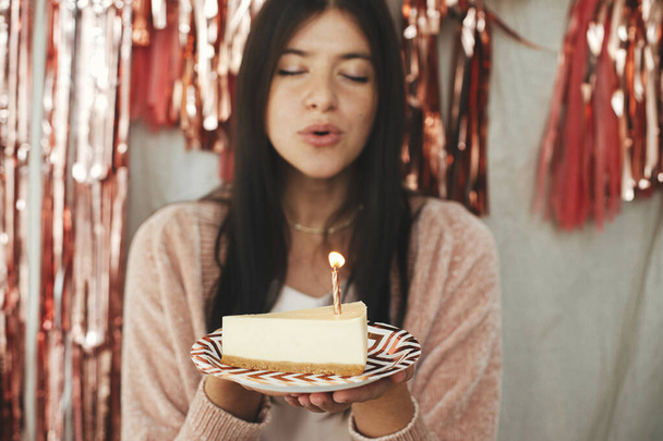 Doe een wens. Stijlvolle gelukkige vrouw blazen brandende kaars op stuk van verjaardagstaart op de achtergrond van moderne rose goud kwastje slinger in de kamer. Vieren verjaardag thuis - Foto, afbeelding