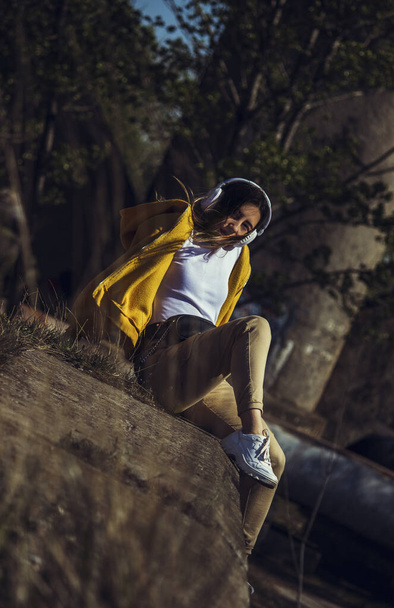 Una chica en un suéter amarillo está sentada en el borde, escuchando música con auriculares blancos y mirando a la cámara. - Foto, imagen