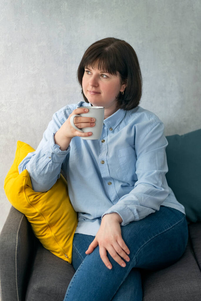 Pensive young woman is holding mug of coffee and sitting on sofa in living room. Coffee break. Vertical frame. - Fotó, kép