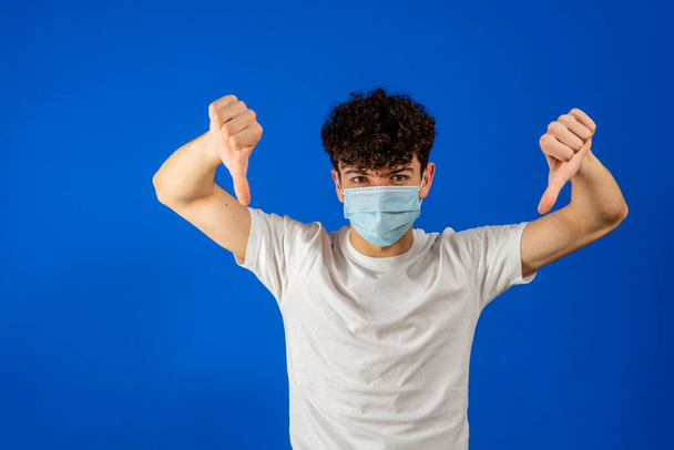 Jeune homme mécontent 20 ans, masque facial stérile à l'abri du coronavirus covid-19, quarantaine pandémique montrer pouce vers le bas détester geste isolé sur fond bleu portrait studio - Photo, image