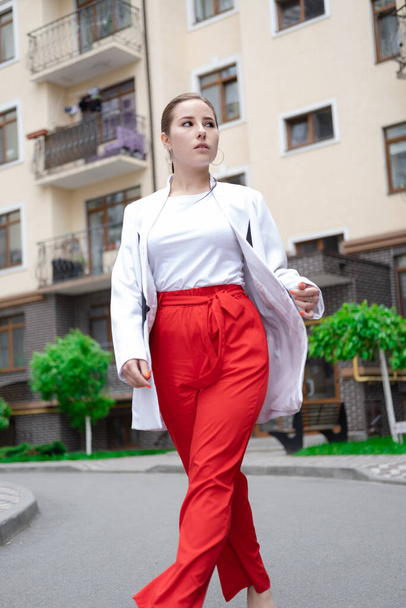pewny brunetka kobieta w czerwony spodnie i biały bluzka i kurtka chodzenie na ulicy. - Zdjęcie, obraz