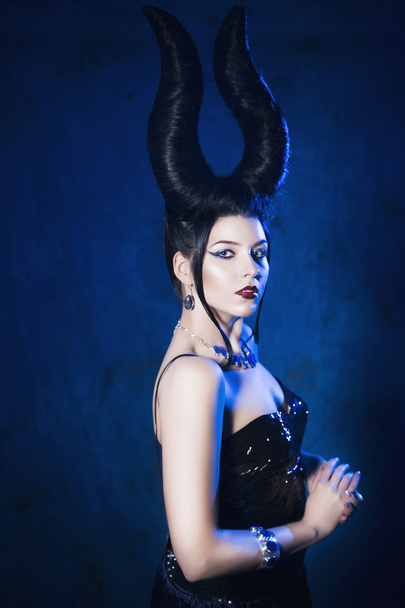 Beautiful woman from a fairytale with a hair horns and jewelry in a spotlight with blue backlight looking at camera - Photo, Image