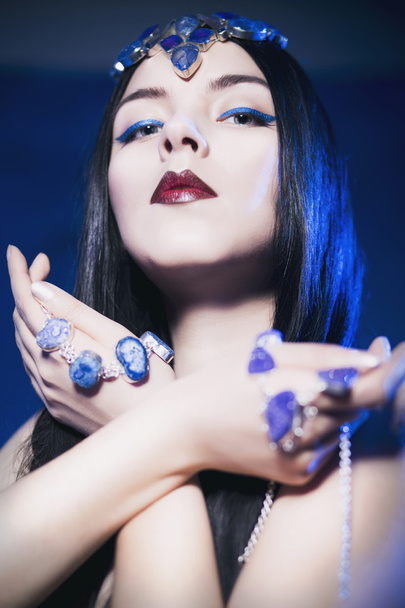 mooie vrouw uit een sprookje met sieraden op haar haar en handen in een schijnwerpers met blauwe achtergrondverlichting camera kijken - Foto, afbeelding