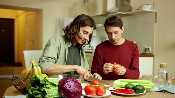 A young man is cutting tomatoes for a salad and talking to his friend - Photo, image