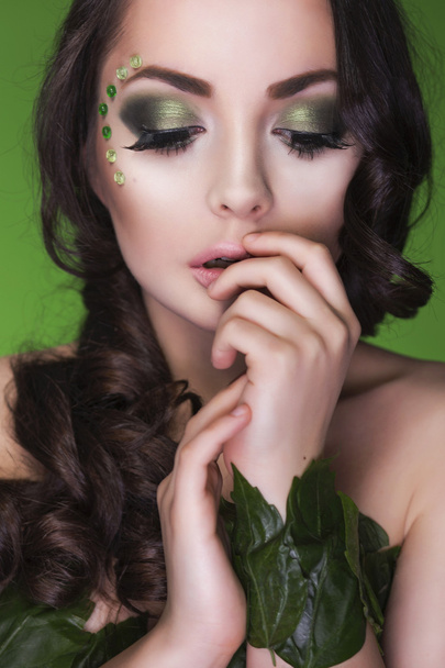mooie brunette dryad vrouw met creatieve make-up en parels op haar gezicht, krullend haar en kostuum gemaakt van bladeren op groene achtergrond. ogen dicht - Foto, afbeelding