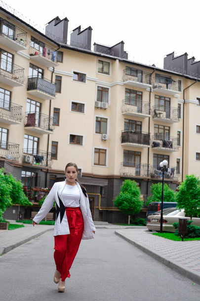 zelfverzekerde brunette vrouw in rode broek en witte blouse en jas lopen in de straat. - Foto, afbeelding