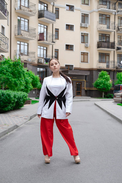 femme brune confiante en pantalon rouge et chemisier blanc et veste marchant dans la rue. - Photo, image