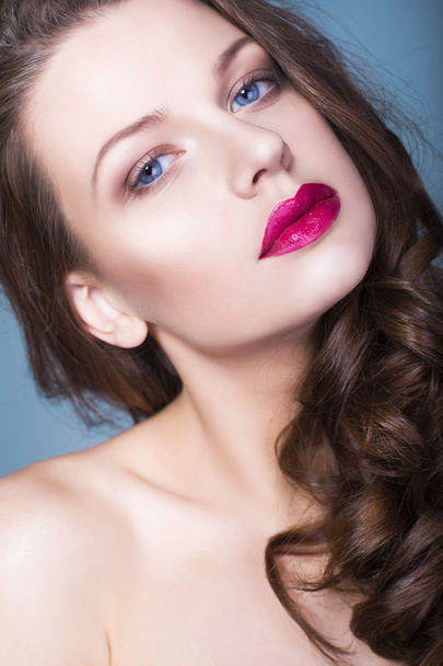 Beautiful brunette woman with creative make up violet eye shadows full red lips, blue eyes and curly hair with her hand on her face - Photo, Image
