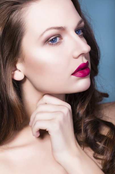 mooie brunette vrouw met creatieve make-up violet eye shadows volledige rode lippen, blauwe ogen en krullend haar met haar hand op haar gezicht - Foto, afbeelding