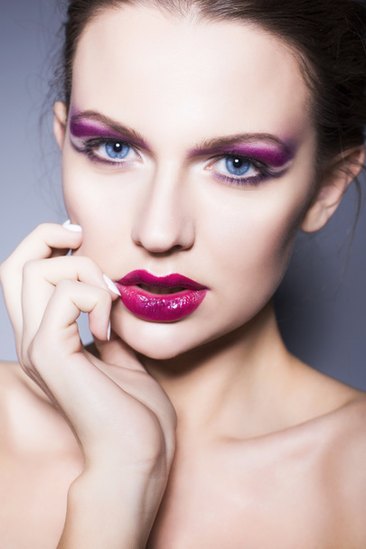 Красивая брюнетка женщина с творческим макияжем фиолетовые тени глаза полные красные губы, голубые глаза и вьющиеся волосы с рукой на лице
 - Фото, изображение