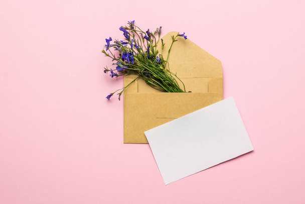 Egy csokor virág egy borítékban, és egy darab fehér papír rózsaszín háttérrel. A szerelmi levelezés fogalma. Lapos fektetés. - Fotó, kép
