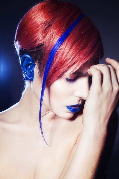 Jeune avec des ombres à paupières bleues, oreilles bleues et cheveux roses avec brin bleu sur elle regardant la caméra et les mains près du visage. Fond noir
 - Photo, image