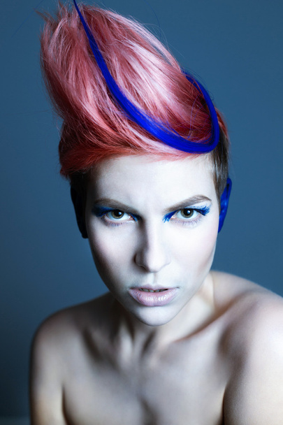 Young person with blue eye shadows, blue ears and pink hair with blue strand on it looking at camera and hands near face. Black background - Foto, Bild