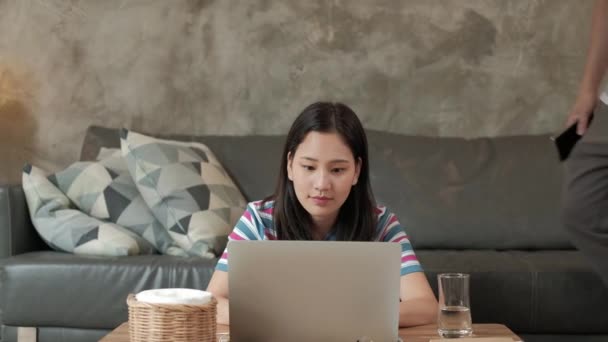 Mujer asiática que utiliza el ordenador portátil para negocios en línea, chat de comercio electrónico, y el aprendizaje de Internet, su hermano en un sofá en la parte posterior. Ambos hermanos trabajan en casa porque tienen que poner en cuarentena al COVID-19. - Imágenes, Vídeo