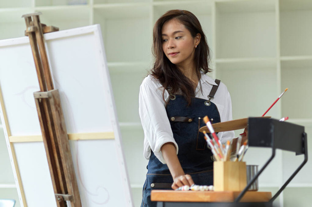 Talented asian female artist painting oil colour on canvas poster, holding palette and paintbrush in studio, creative occupation - Zdjęcie, obraz