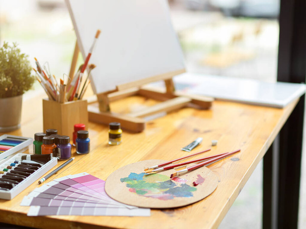 Primer plano, espacio de trabajo del artista con caballete de tela, paleta, pinceles, muestra de muestras de muestras de color, lápices de colores pastel, acrílico sobre mesa de madera - Foto, imagen